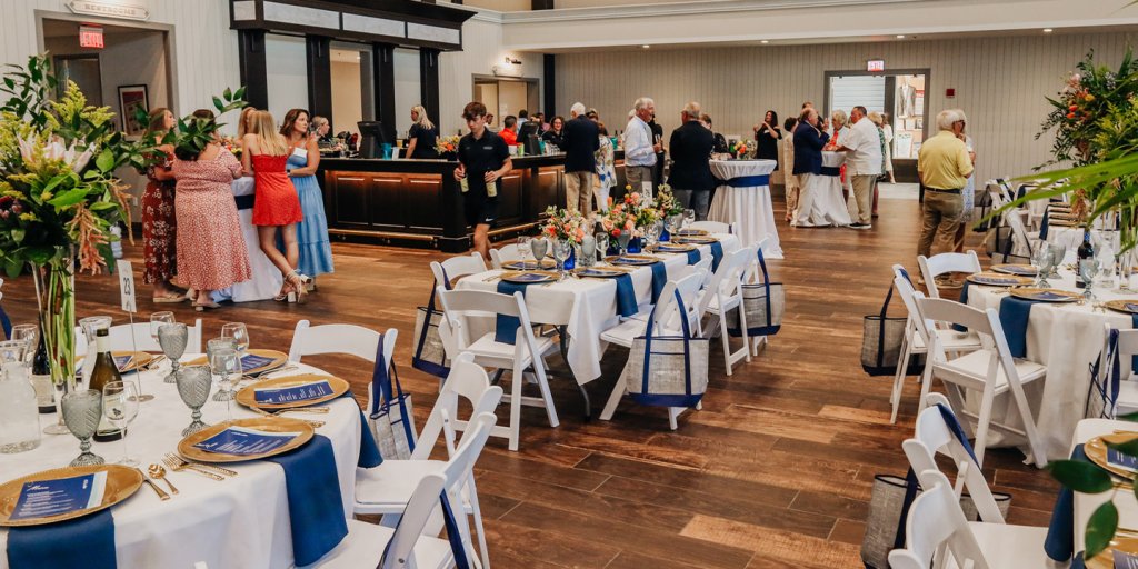 A group of people at an event surround a bar and cocktail tables. with more tables and chairs set for dinner.