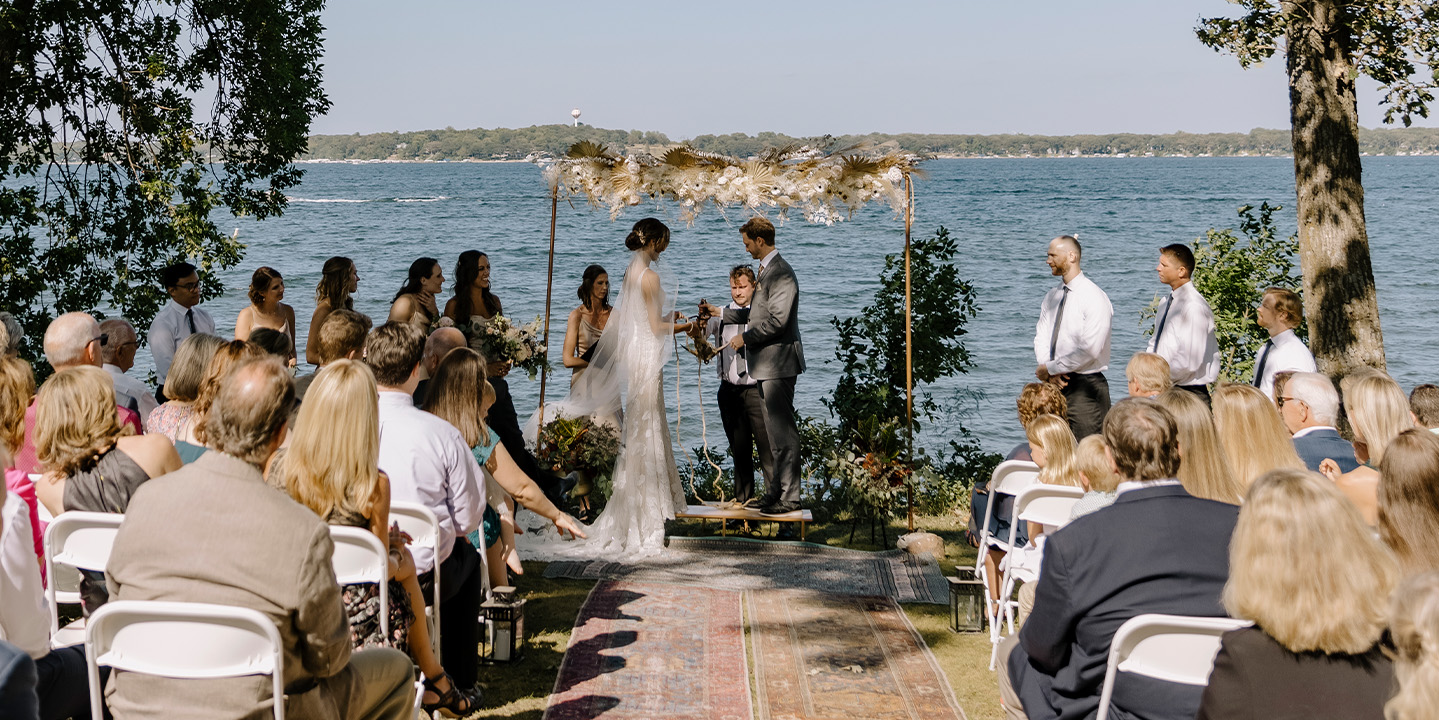 Weddings 2 A bride and groom get married under an arch lakeside with family and friends in attendance.